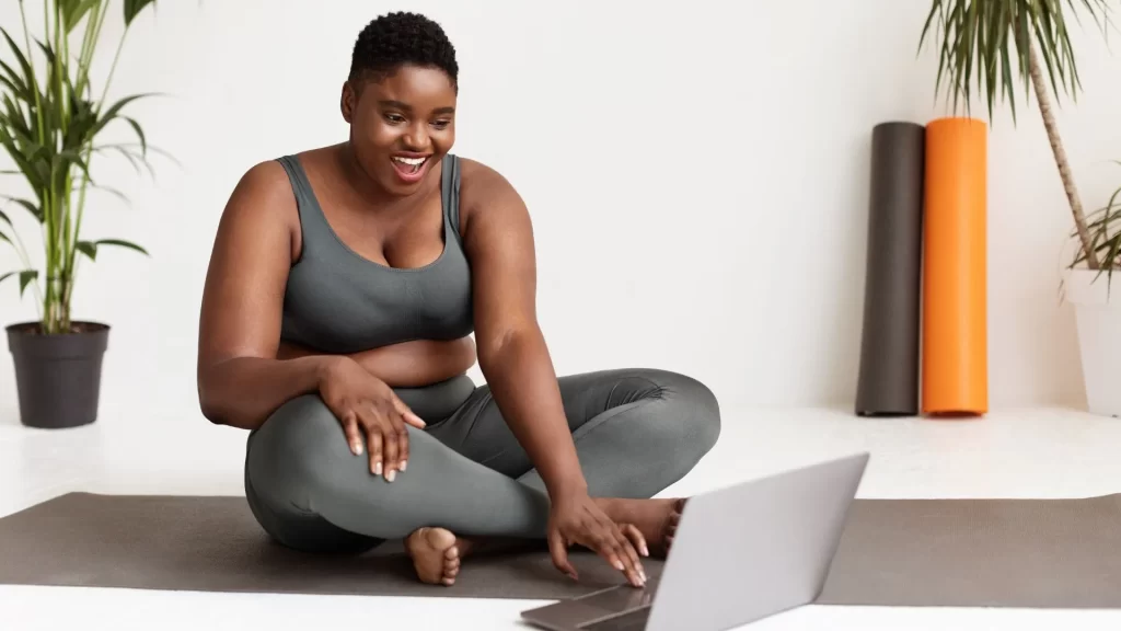 An African American woman in gym wear, sitting in a yoga mat while happily using her laptop