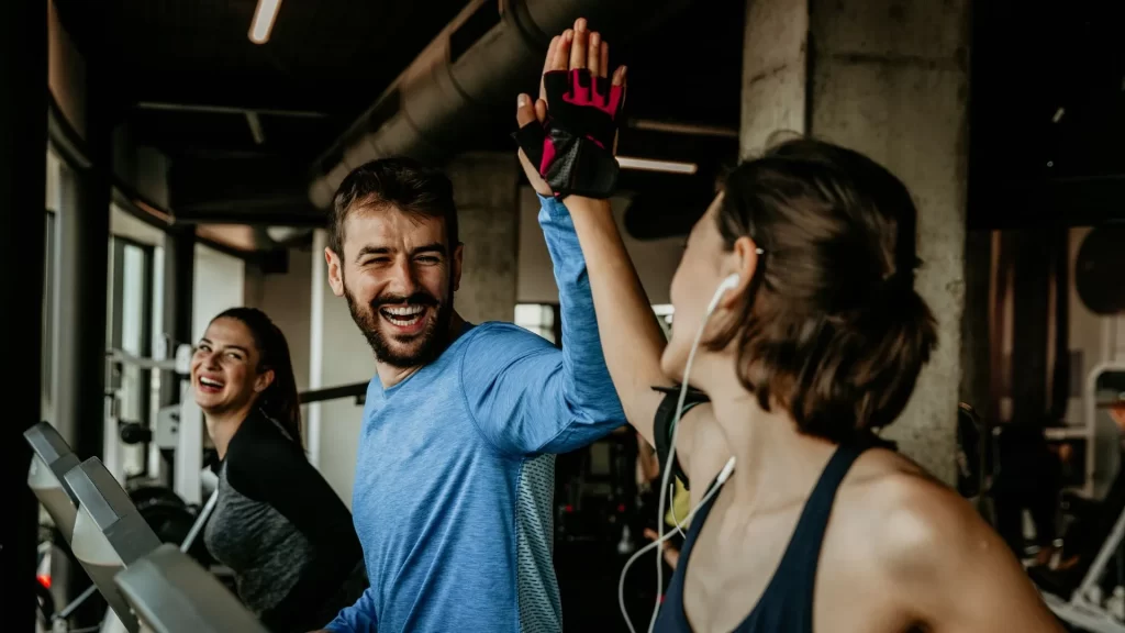 A man and a woman do a high five while running in a treadmill as their other woman friend smiles from the side