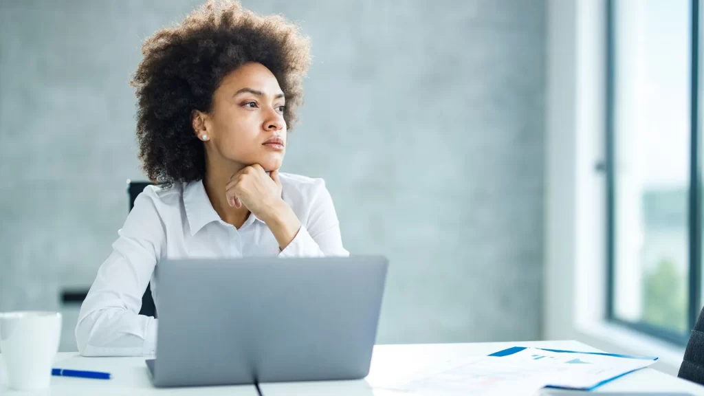 A woman in corporate attire is staring into space lost in her thoughts while her laptop is open in front of her