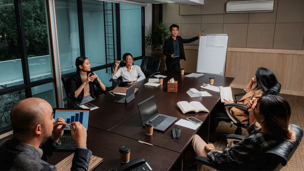 An Asian man is happily presenting something in a small office conference room. His colleagues are listening intently while their laptops and work essentials are all on the table. 