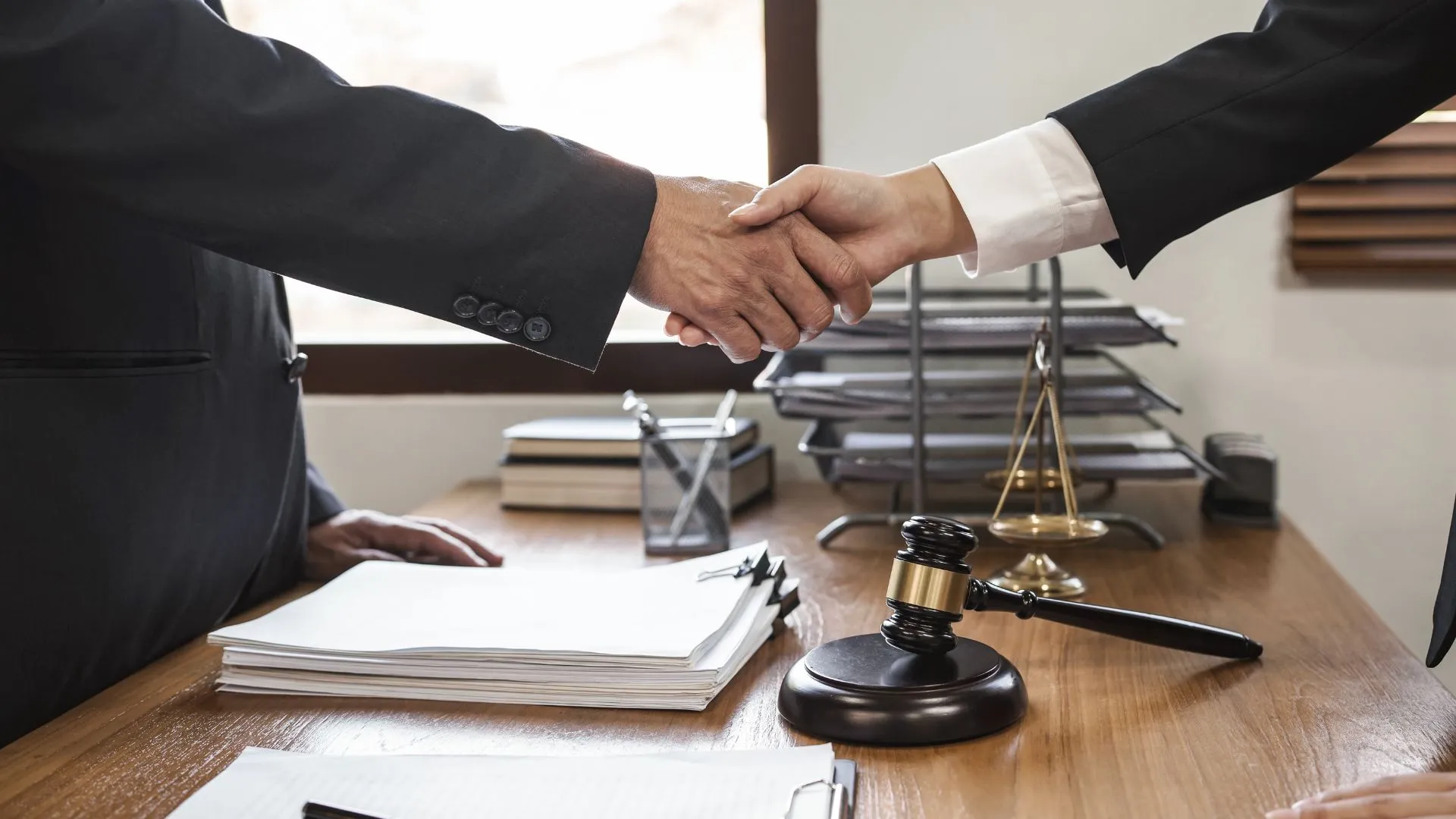 Two lawyers shake hands over a wooden office table. A justice scale and a gavel decorate the table.