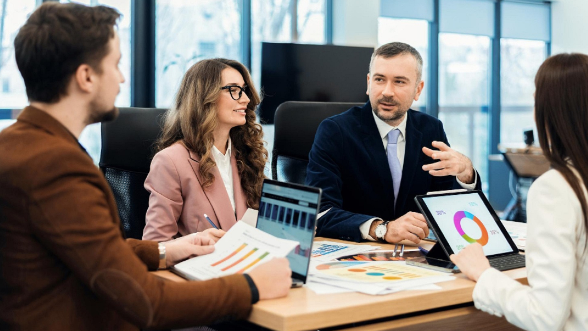 An image depicts four professionals around a conference table in an office setting. Two men and two women are shown.